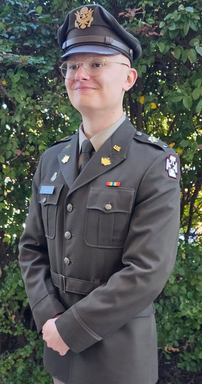 Smiling young man in a US Army dress uniform and service cap standing in front of greenery