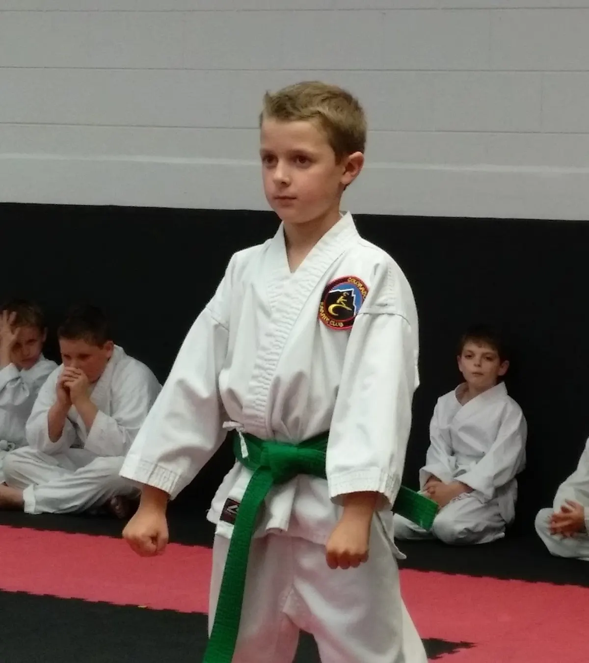 Young karate student with a green belt standing at attention while classmates watch from the side of the mat