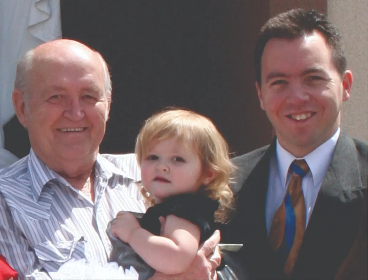 Older man and younger man in a suit smiling with a toddler girl held between them